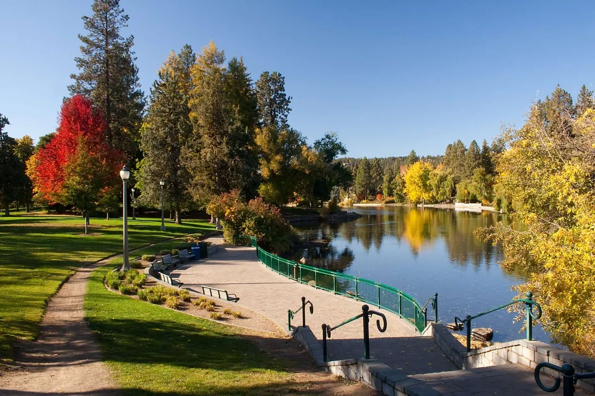 A serene lake scene bordered by colorful autumn trees, a winding walking path, and park benches perfect for relaxation. The tranquil mood invites walking, picnicking, or quiet moments by the water.