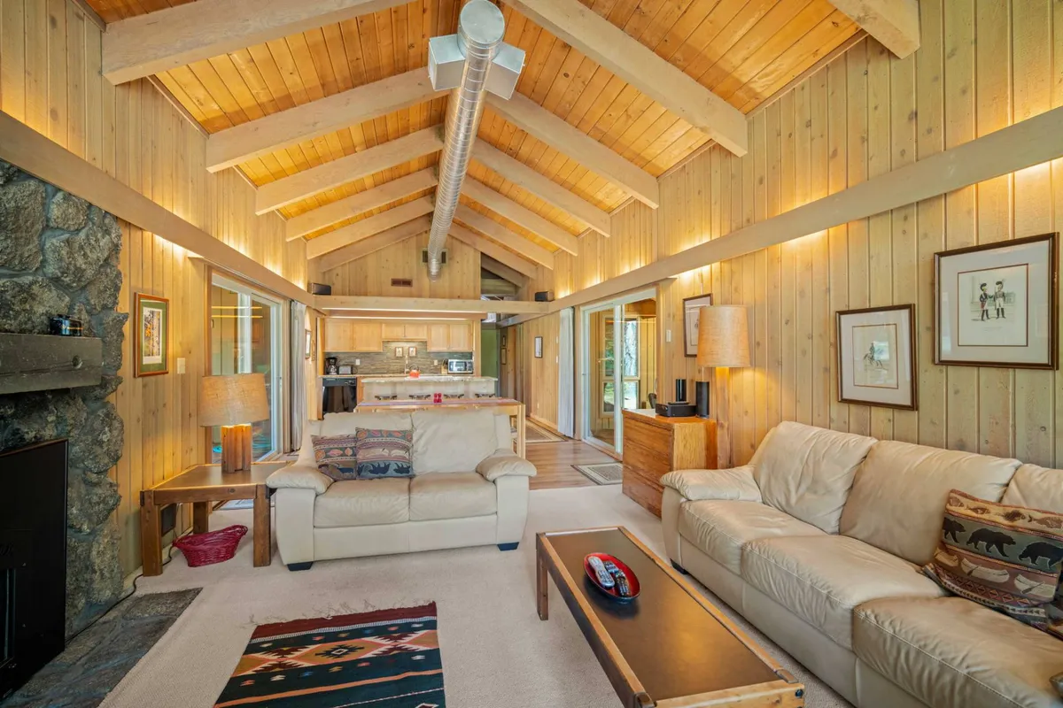 This image showcases a spacious living area featuring a dramatic stone fireplace, large windows flooding the space with natural light, high wooden ceilings, comfortable sofas, and a decorative rug. The layout encourages relaxation and togetherness, with ample seating and warm, welcoming decor.