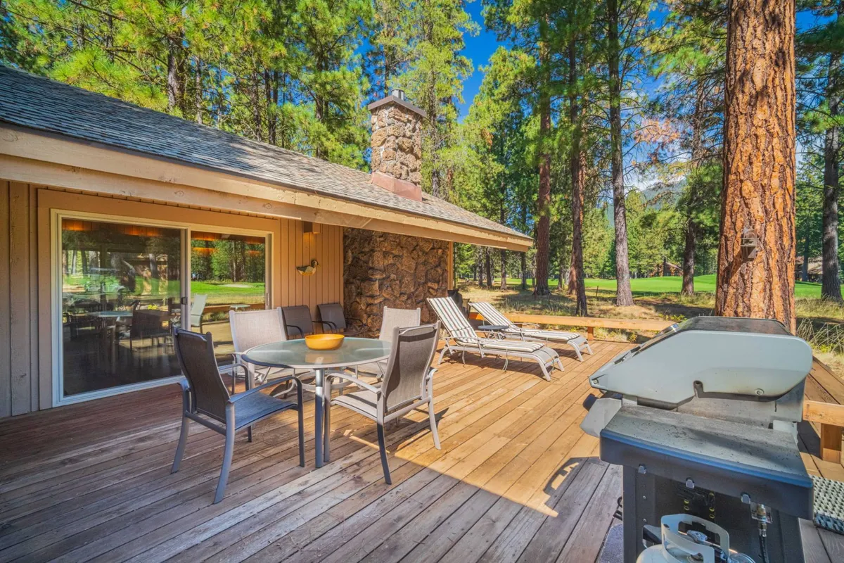 This image shows a spacious outdoor patio featuring a table with chairs, a gas grill, and lounge seating, all arranged around a striking stone chimney. The area is bathed in warm, natural light and surrounded by lush greenery, perfectly capturing the ambiance of outdoor relaxation and al fresco dining.