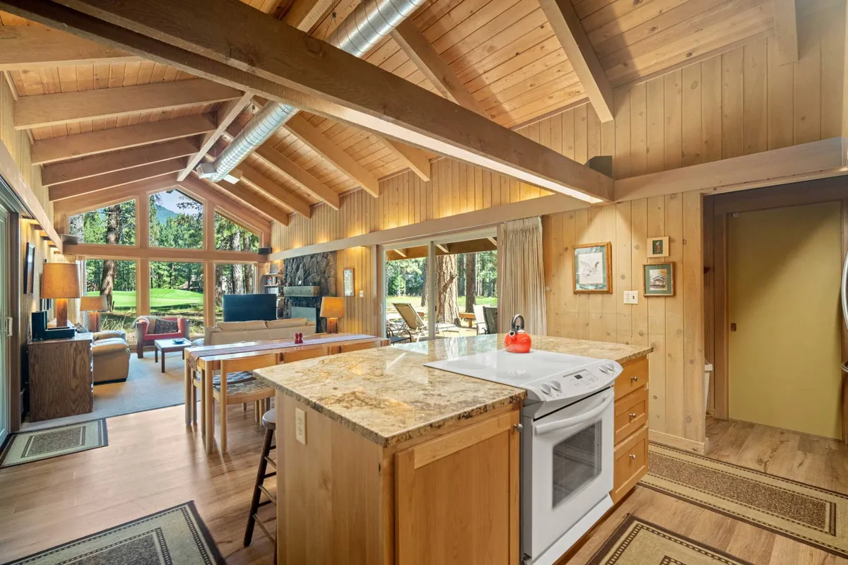 A view of the open-concept kitchen and adjoining living space, featuring a granite kitchen island, large windows with views of the outdoors, wooden beams, and a comfortable seating area. The scene is bright and airy, highlighting seamless flow between kitchen, dining, and living zones.