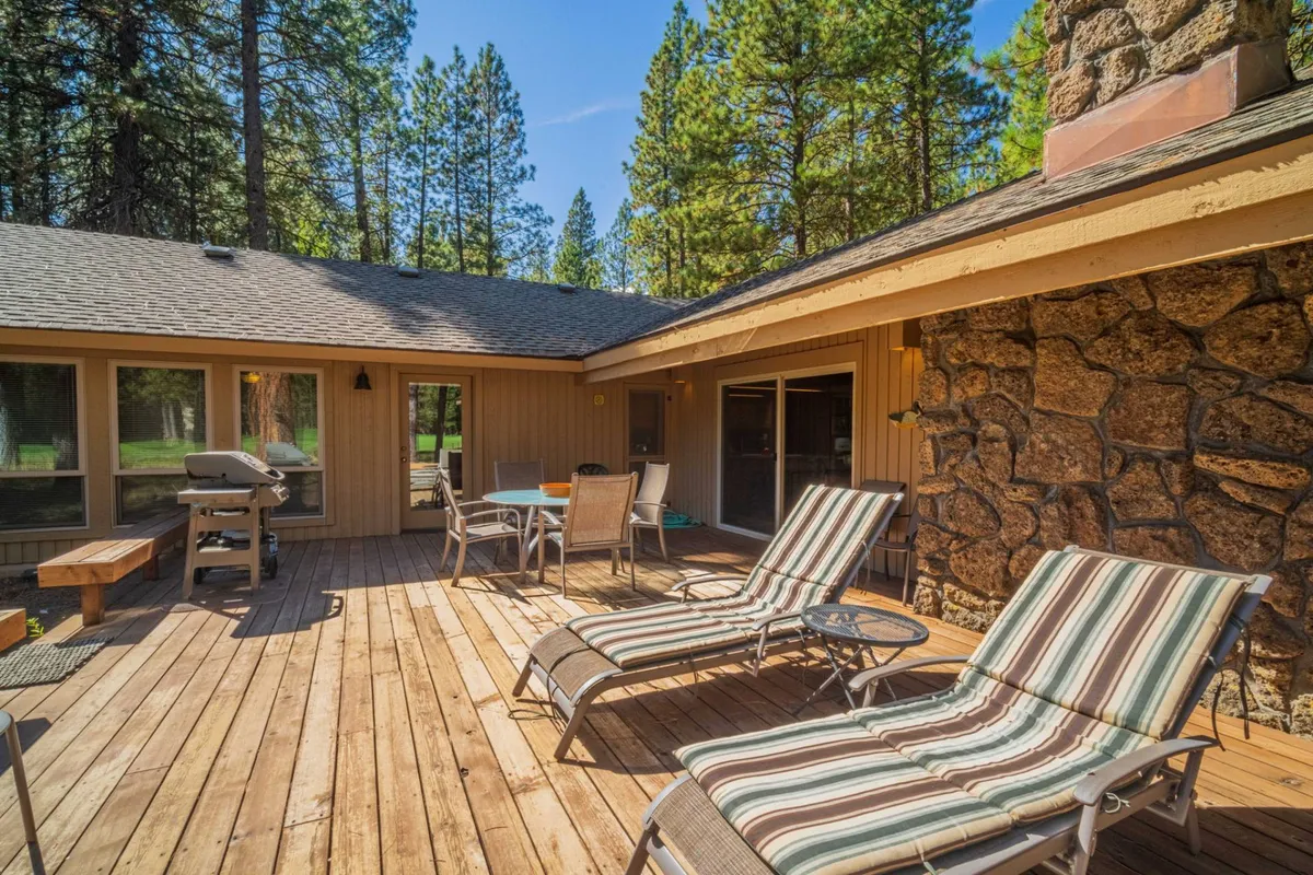 This image features a deck with lounge chairs, a dining table, a gas grill, and a low stone wall, all set against a backdrop of trees. The scene suggests both relaxation and social activities, such as sunbathing, grilling, or enjoying meals outside.