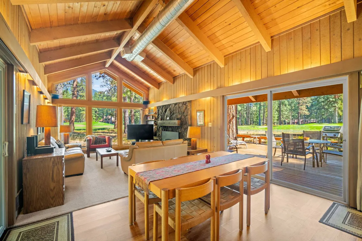 A bright, open-concept living area featuring large windows, a dining table, a cozy living room with a fireplace, and direct access to the outdoor deck. The space is arranged to foster group interaction and offers beautiful views of the surrounding landscape, ideal for shared meals and game nights.