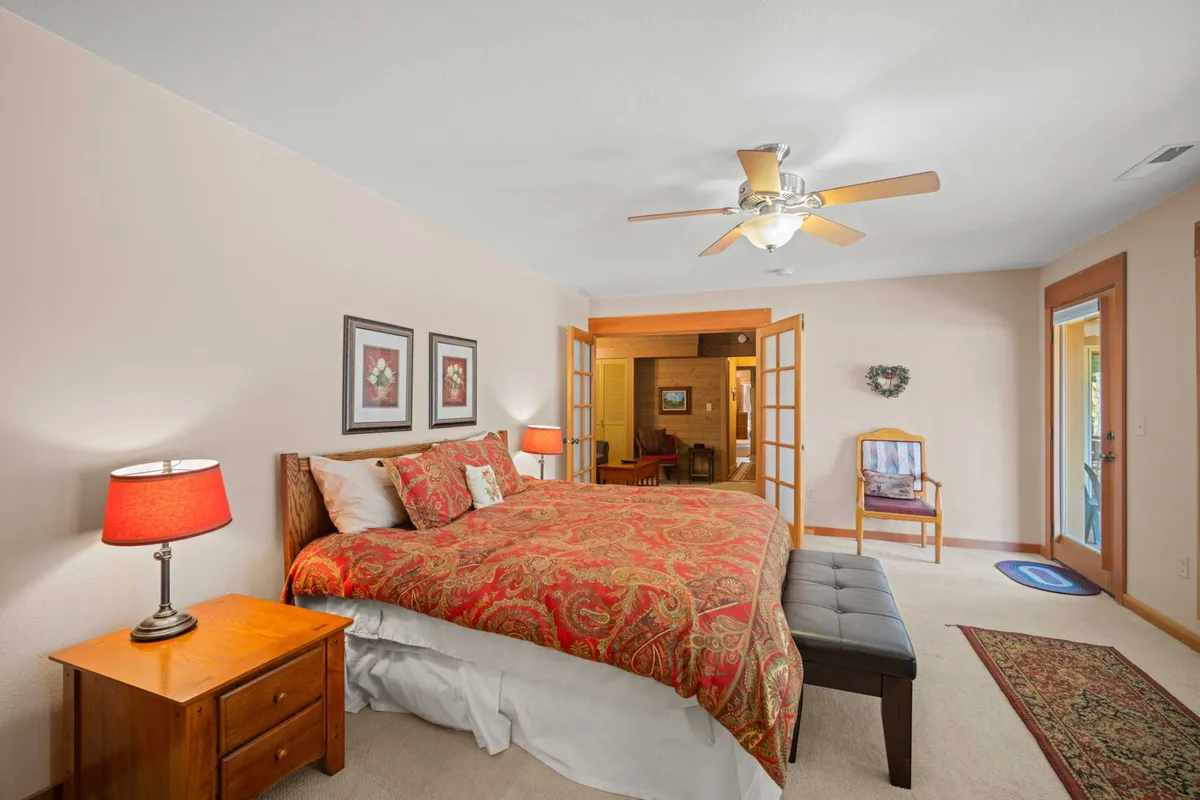 A spacious bedroom featuring a large bed with ornate bedding, a nightstand with a lamp, French doors leading to another room, and a decorative chair. The room is bathed in soft natural light, creating a serene and welcoming atmosphere ideal for relaxation.