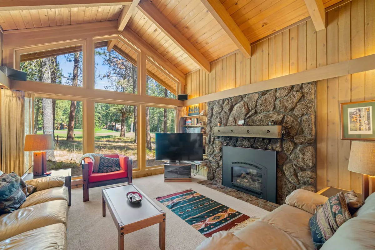 The image showcases a cozy, sunlit living area with expansive windows framing tranquil forest views, a striking stone fireplace, and plush seating arranged on a decorative rug, all creating a welcoming space for relaxation and togetherness.