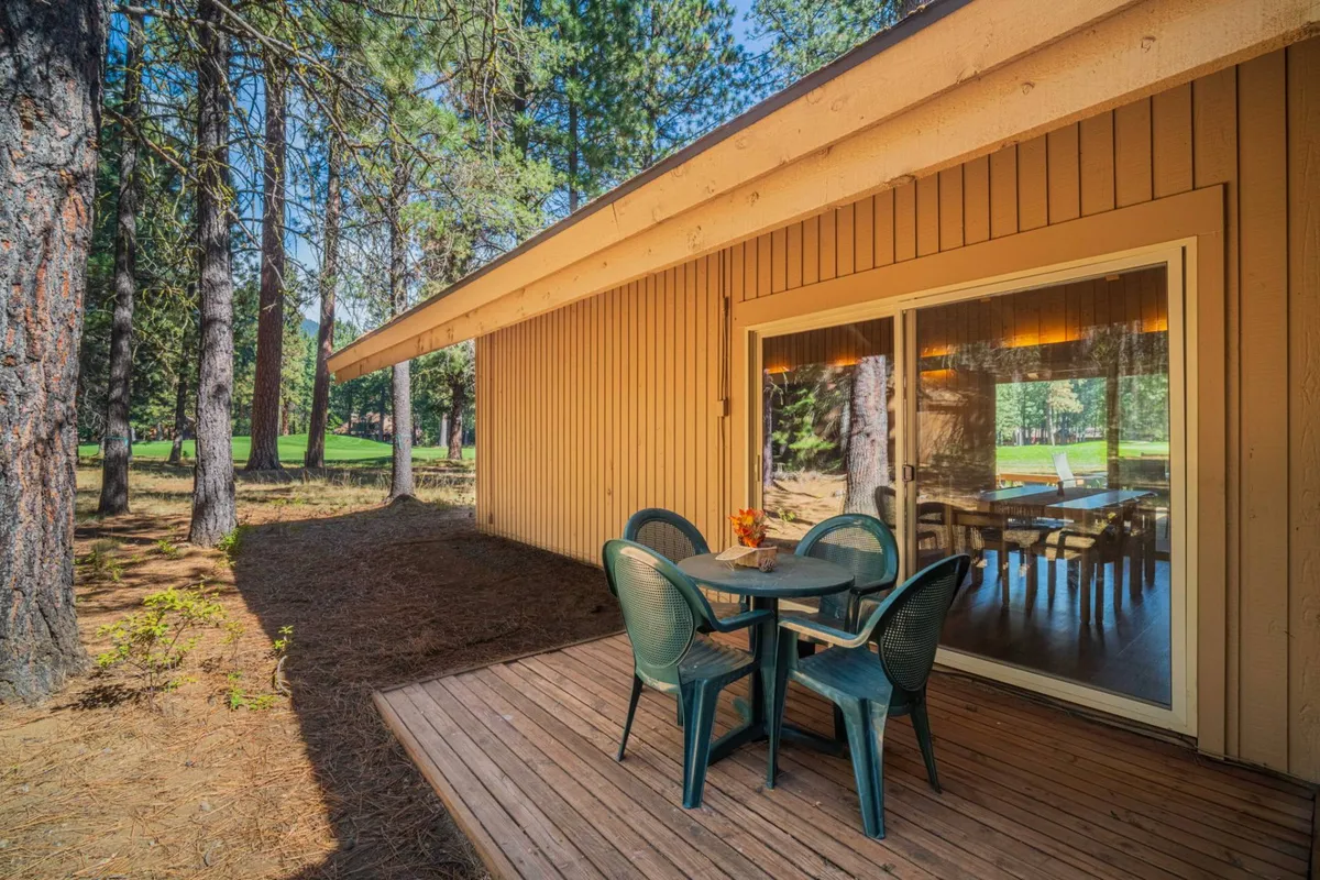 An inviting outdoor dining area set beneath tall trees, with a large wooden table surrounded by chairs. The space is bathed in soft, natural light filtering through the canopy, and large glass doors connect the area seamlessly to the interior. The tranquil setting is ideal for enjoying morning coffee, alfresco meals, or simply relaxing while surrounded by nature.