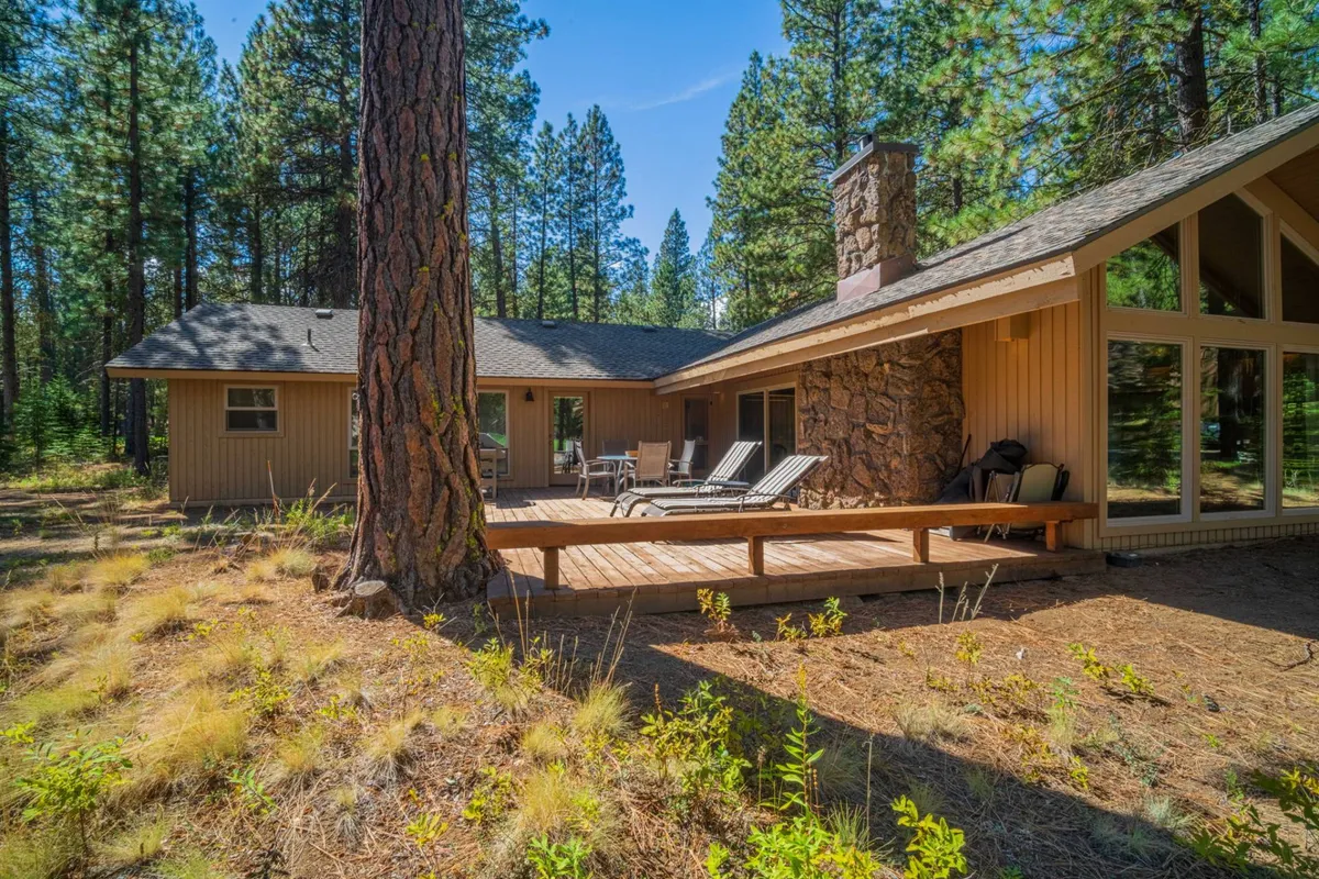 A peaceful exterior shot of a wooden deck surrounded by tall trees, with large windows and a stone chimney hinting at the cozy home inside. The deck offers space for outdoor dining, relaxing, or taking in nature views.