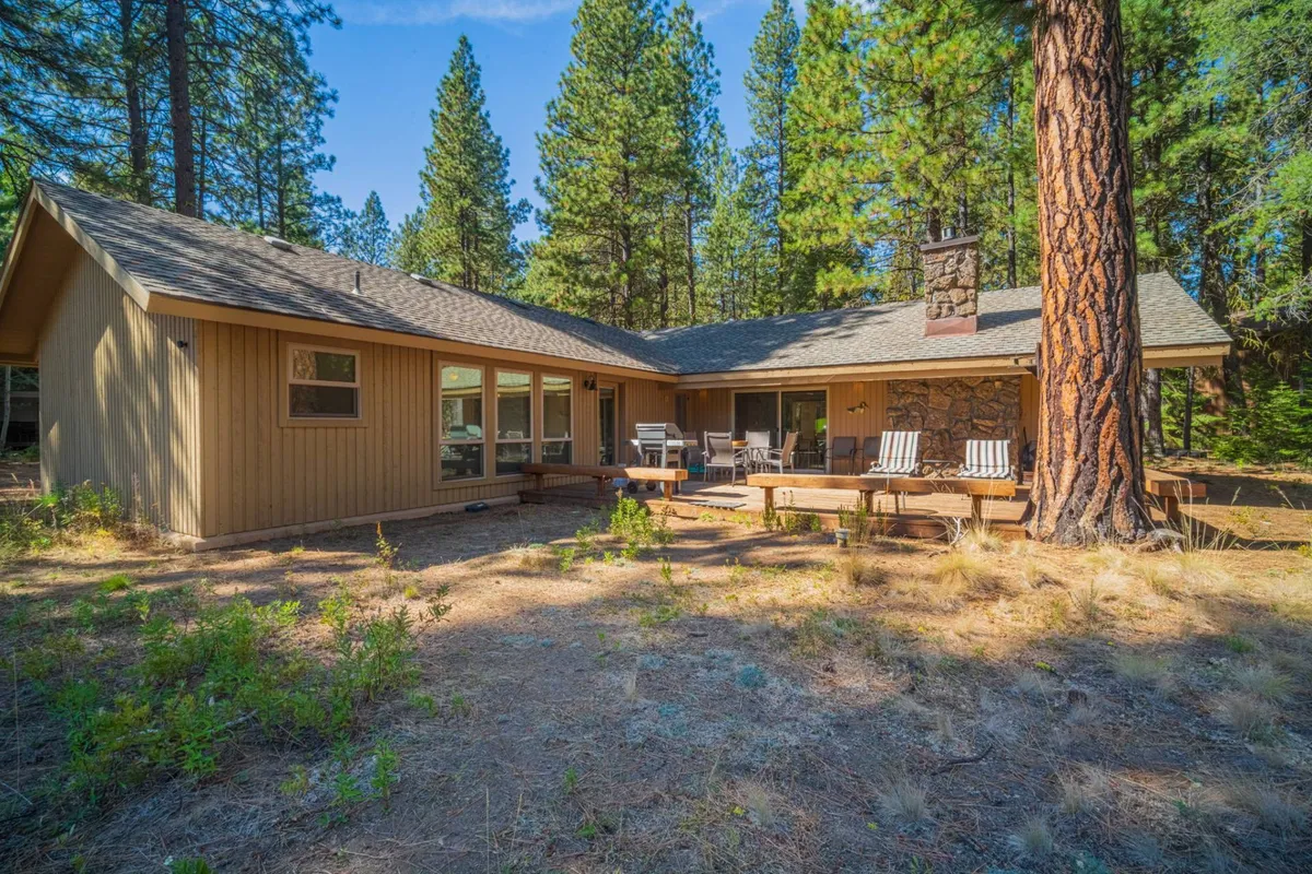 A rustic wooden deck attached to a standalone home, highlighted by a charming stone chimney and framed by tall forest trees. The deck offers space for seating and relaxation, surrounded by the peace and beauty of nature. The scene captures either the golden hour or early evening, hinting at sunset views and the potential for stargazing at night.