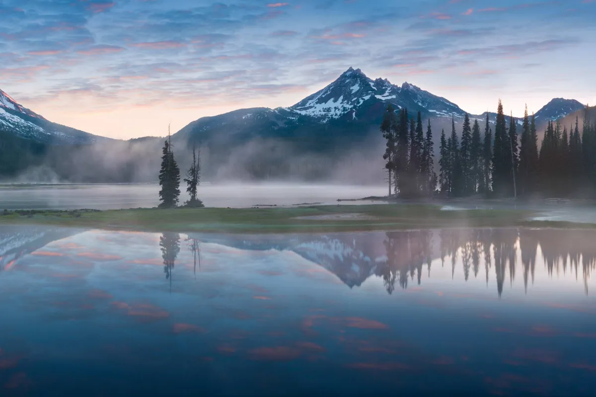 A breathtaking view of a calm lake bordered by tall evergreen trees, with snow-capped mountains rising in the distance and a soft mist hovering over the water. The scene is tranquil, natural, and feels inviting for hiking, fishing, and nature walks.